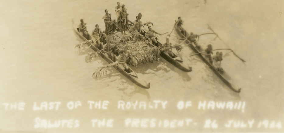 Outrigger canoes, one with a platform covered in what are maybe ki leaves bearing a man in a traditional feathered cape and headdress. Click to enlarge.